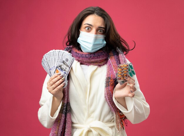 Impressionato giovane donna malata che indossa accappatoio e sciarpa con maschera che tiene soldi e confezioni di capsule mediche guardando davanti isolato sul muro rosa