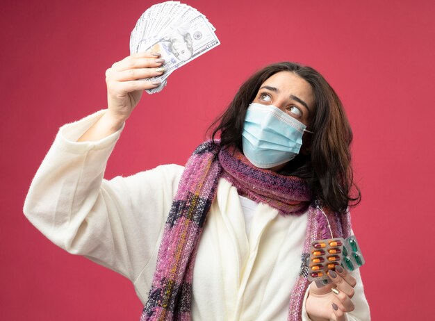 Impressionato giovane donna malata che indossa accappatoio e sciarpa con maschera che tiene soldi e confezioni di capsule mediche sollevando e guardando soldi isolati sul muro rosa