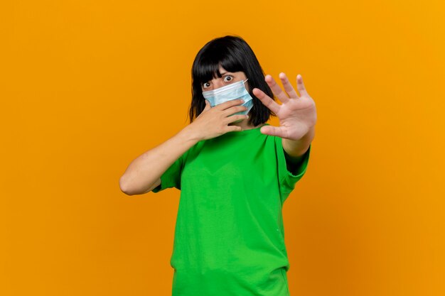 Impressionato giovane donna malata che indossa la maschera toccando il viso allungando la mano verso la parte anteriore guardando la parte anteriore isolata sulla parete arancione