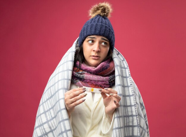 Impressionato giovane donna malata che indossa un abito invernale cappello e sciarpa avvolti in plaid che tiene intonaco medico guardando il lato isolato sul muro rosa