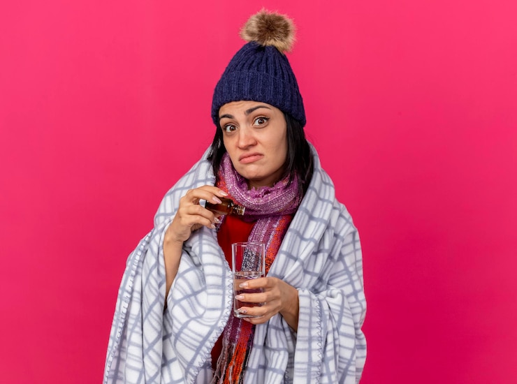 Foto gratuito impressionato giovane donna malata che indossa un cappello invernale e sciarpa avvolta in un plaid aggiungendo medicamento nel bicchiere d'acqua guardando la parte anteriore isolata sul muro rosa con spazio di copia