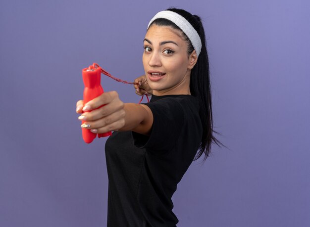 Impressionato giovane donna sportiva che indossa fascia e braccialetti in piedi in vista di profilo tirando la corda per saltare allungandola verso la parte anteriore guardando la parte anteriore isolata sul muro viola