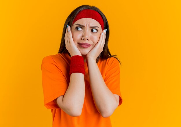 Impressionato giovane donna sportiva che indossa la fascia e braccialetti mantenendo le mani sul viso guardando a lato