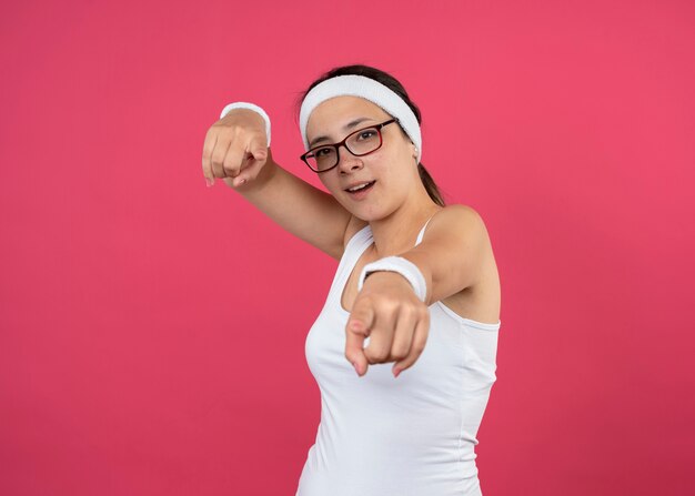 Impressionato giovane donna sportiva con occhiali ottici che indossa fascia e braccialetti che punta sul davanti isolato sul muro rosa