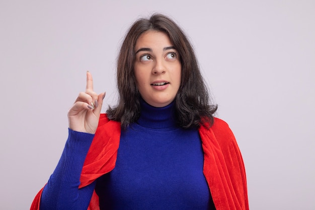 Impressionato giovane donna supereroe in mantello rosso che guarda il lato rivolto verso l'alto isolato sul muro bianco