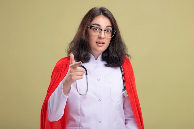 Impressionato giovane donna supereroe in mantello rosso che indossa l'uniforme del medico e lo stetoscopio con gli occhiali che guardano e puntano sul davanti isolato sul muro verde oliva con spazio di copia