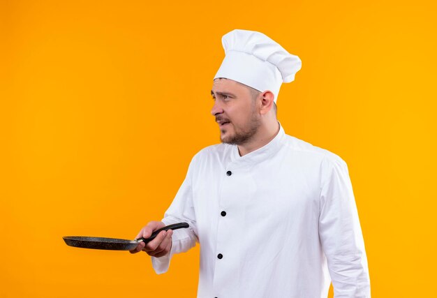 Impressionato giovane e bello cuoco in uniforme da chef che tiene in mano una padella che guarda il lato isolato sulla parete arancione