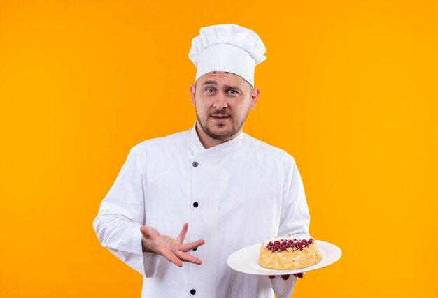 Impressionato giovane e bello cuoco in uniforme da chef che tiene un piatto di torta e lo indica isolato su una parete arancione