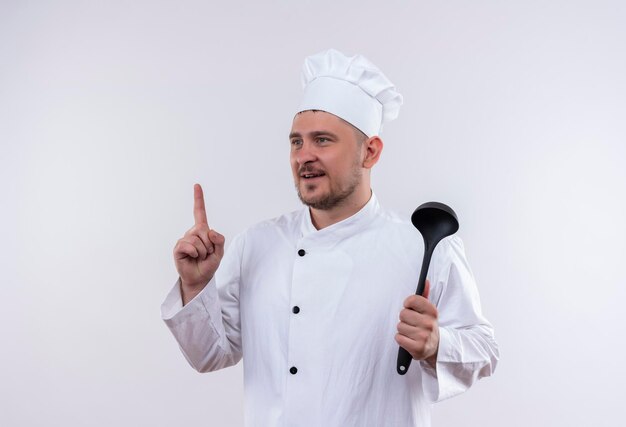 Impressionato giovane e bello cuoco in uniforme da chef tenendo il mestolo e alzando il dito guardando il lato isolato sul muro bianco