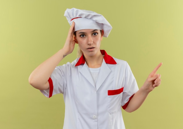 Impressionato giovane e graziosa cuoca in uniforme da chef mettendo la mano sulla testa e indicando il lato isolato sul muro verde