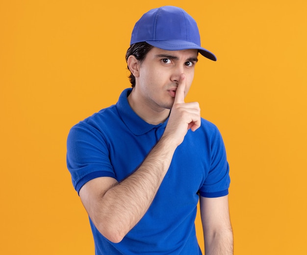 Impressionato giovane fattorino caucasico in uniforme blu e berretto che fa gesto di silenzio solato su sfondo arancione
