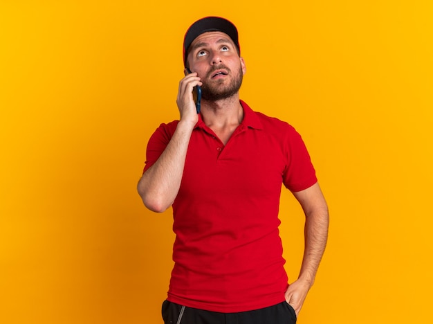 Impressionato giovane fattorino caucasico in uniforme rossa e berretto che tiene la mano in tasca parlando al telefono guardando in alto isolato sulla parete arancione con spazio di copia