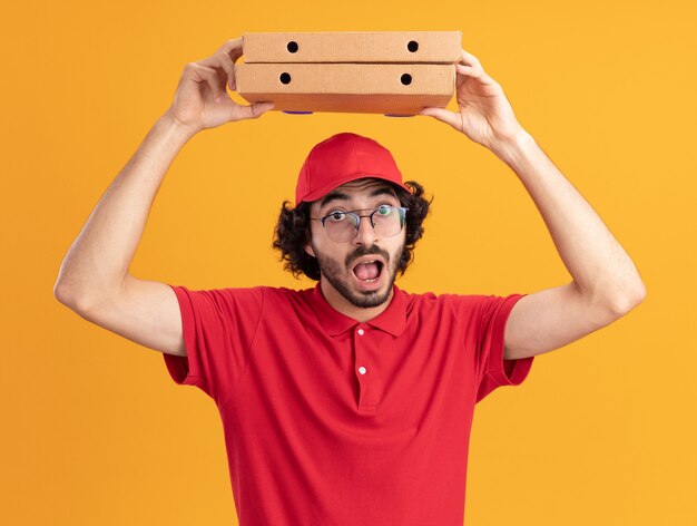 Impressionato giovane fattorino caucasico in uniforme rossa e berretto con gli occhiali che tengono i pacchetti di pizza sopra la testa