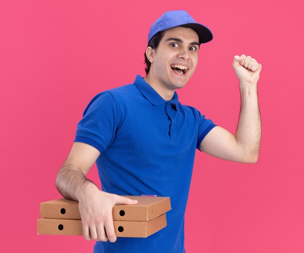 Impressionato giovane fattorino in uniforme blu e berretto in piedi in vista di profilo con in mano pacchetti di pizza facendo gesto di bussare guardando la parte anteriore isolata sul muro rosa