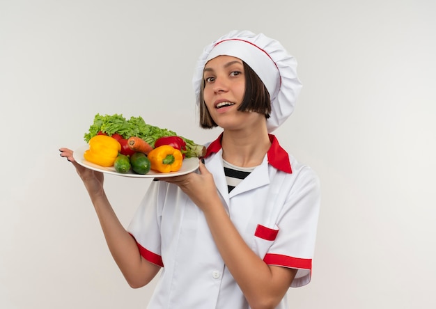 Impressionato giovane femmina cuoco in chef uniforme tenendo piatto di verdure isolato su bianco con copia spazio