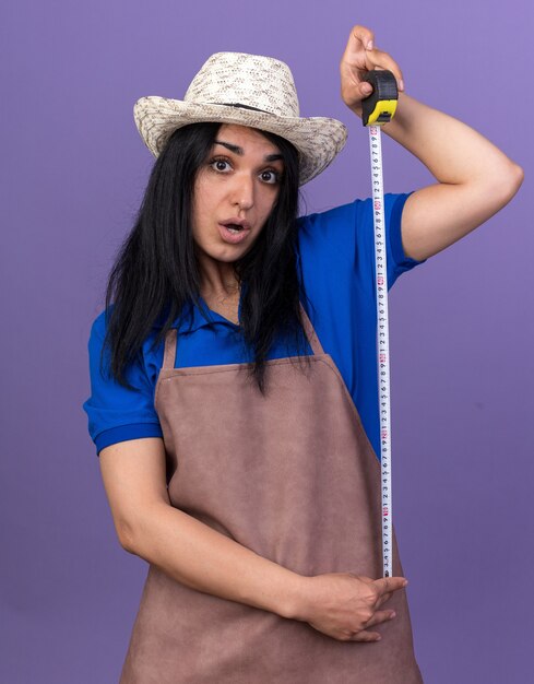 Impressionato giovane giardiniere donna che indossa l'uniforme e cappello tenendo il metro a nastro guardando la parte anteriore isolata sul muro viola