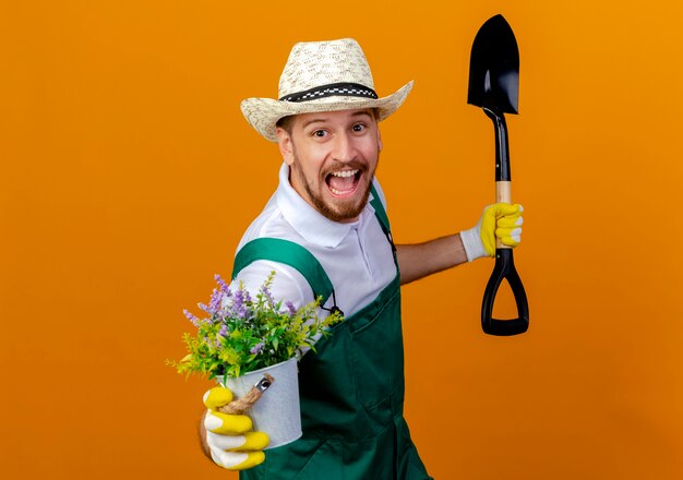 Impressionato giovane giardiniere slavo bello in uniforme che indossa cappello e guanti da giardinaggio che tengono la vanga e allungando il vaso di fiori isolato