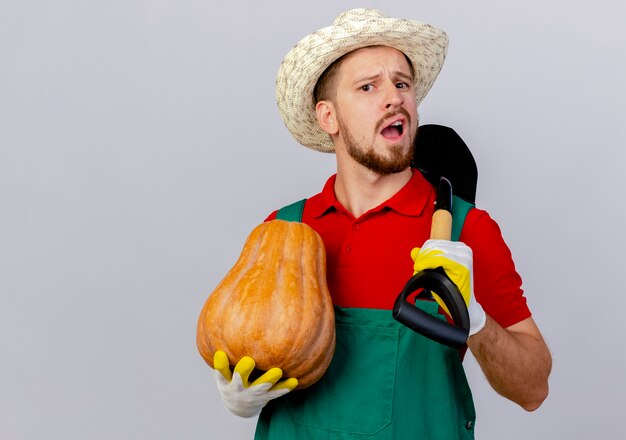 Impressionato giovane giardiniere slavo bello in uniforme che indossa guanti da giardinaggio e cappello che tiene zucca butternut e vanga cercando isolato