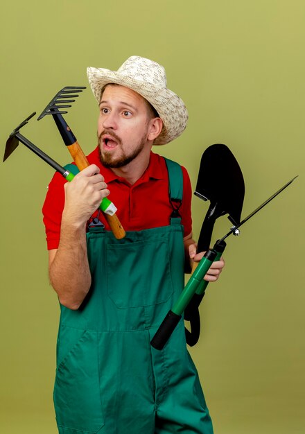 Impressionato giovane giardiniere slavo bello in uniforme e cappello che tiene e guardando gli strumenti del giardiniere