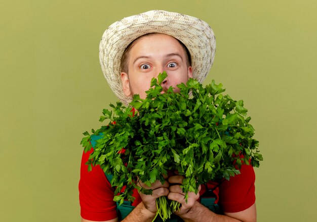 Impressionato giovane giardiniere slavo bello in uniforme e cappello che tiene il coriandolo da dietro isolato sulla parete verde oliva