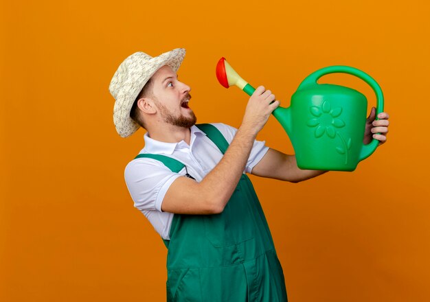 Impressionato giovane giardiniere slavo bello in uniforme e cappello tenendo e guardando annaffiatoio isolato