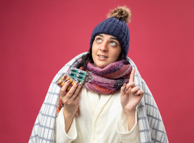 Impressionato giovane indoeuropea ragazza malata che indossa abito invernale cappello e sciarpa avvolti in plaid con confezioni di capsule guardando e rivolto verso l'alto isolato sul muro cremisi