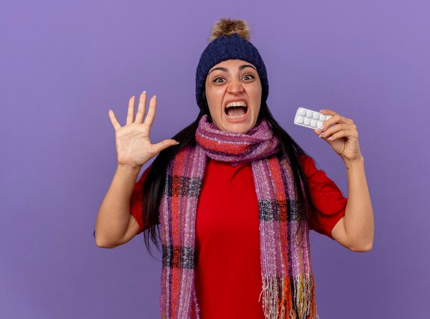 Impressionato giovane indoeuropea ragazza malata che indossa un cappello invernale e sciarpa che mostra confezione di compresse guardando la telecamera che mostra cinque con la mano isolata su sfondo viola