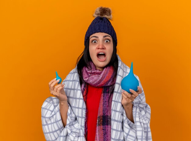 Impressionato giovane indoeuropeo ragazza malata che indossa cappello invernale e sciarpa avvolto in plaid che tiene i clisteri guardando la telecamera isolata su sfondo arancione con spazio di copia