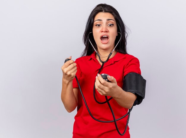 Impressionato giovane indoeuropeo ragazza malata che indossa uno stetoscopio misurando la sua pressione con sfigmomanometro guardandolo isolato su sfondo bianco con spazio di copia