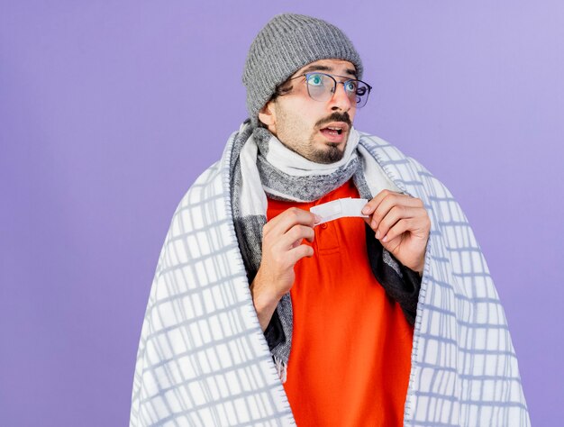 Impressionato giovane indoeuropeo uomo malato con gli occhiali inverno cappello e sciarpa avvolto in plaid guardando lato tenendo intonaco medico isolato su sfondo viola con spazio di copia