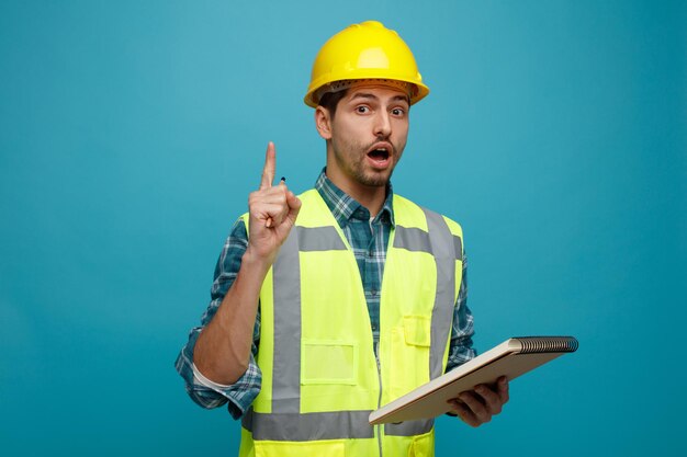 Impressionato giovane ingegnere maschio che indossa casco di sicurezza e uniforme che tiene matita e blocco note guardando la fotocamera rivolta verso l'alto isolata su sfondo blu