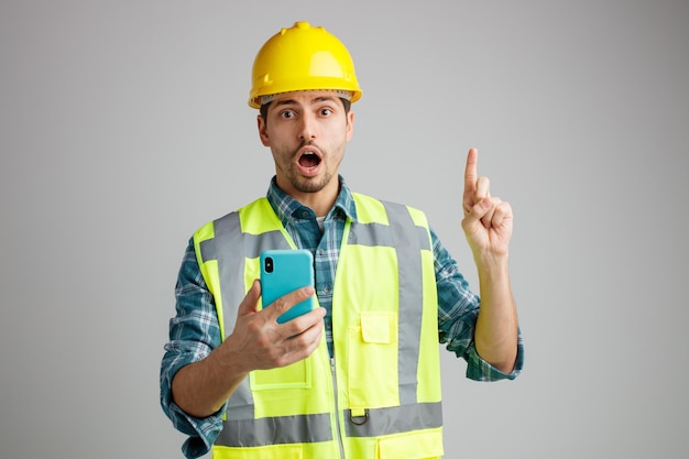 Impressionato giovane ingegnere maschio che indossa un casco di sicurezza e uniforme che tiene il telefono cellulare guardando la fotocamera rivolta verso l'alto isolata su sfondo bianco