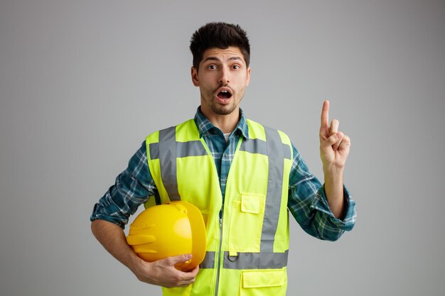Impressionato giovane ingegnere maschio che indossa uniforme che tiene il casco di sicurezza guardando la fotocamera rivolta verso l'alto isolata su sfondo bianco