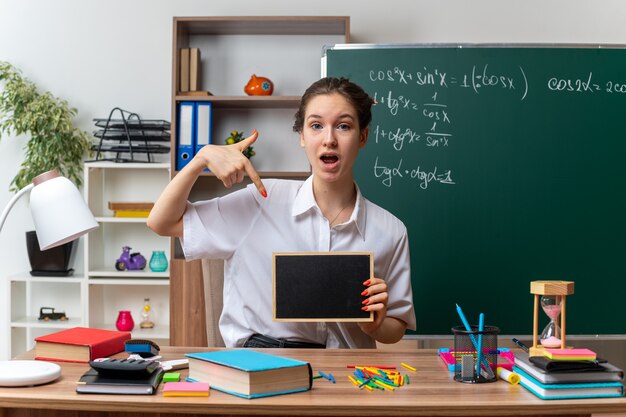 Impressionato giovane insegnante di matematica femminile bionda seduto alla scrivania con strumenti di scuola che tengono e puntano alla mini lavagna guardando la fotocamera in aula