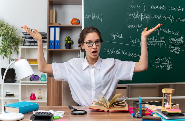 impressionato giovane insegnante di matematica femminile con gli occhiali seduto alla scrivania con materiale scolastico guardando la parte anteriore che mostra le mani vuote in classe