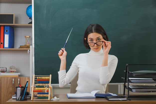 impressionato giovane insegnante femminile che indossa occhiali punti con puntatore alla lavagna seduto alla scrivania con strumenti scolastici in classe