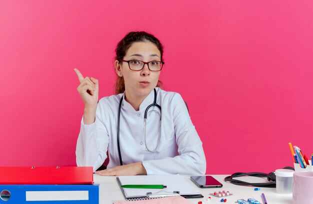 Impressionato giovane medico femminile che indossa abito medico e stetoscopio e occhiali seduto alla scrivania con strumenti medici cercando di alzare il dito isolato