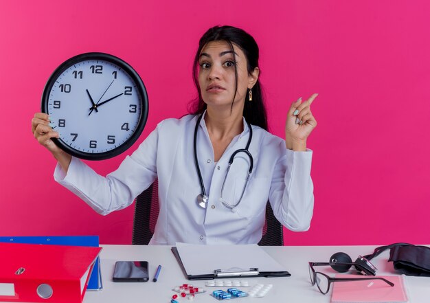 Impressionato giovane medico femminile che indossa abito medico e stetoscopio seduto alla scrivania con strumenti medici che tengono l'orologio e alzando il dito isolato sul muro rosa