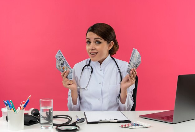 Impressionato giovane medico femminile che indossa una veste medica con lo stetoscopio seduto alla scrivania lavora sul computer con strumenti medici che tengono contanti sulla parete rosa isolata con lo spazio della copia