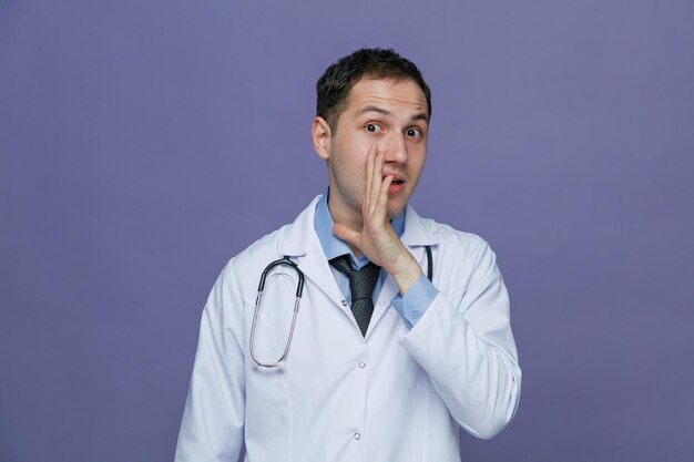 Impressionato giovane medico maschio che indossa accappatoio medico e stetoscopio intorno al collo guardando la fotocamera tenendo la mano vicino alla bocca sussurrando isolato su sfondo viola