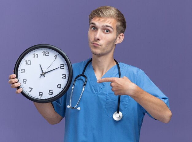 Impressionato giovane medico maschio che indossa l'uniforme del medico con la tenuta dello stetoscopio e punti all'orologio da parete isolato sulla parete blu