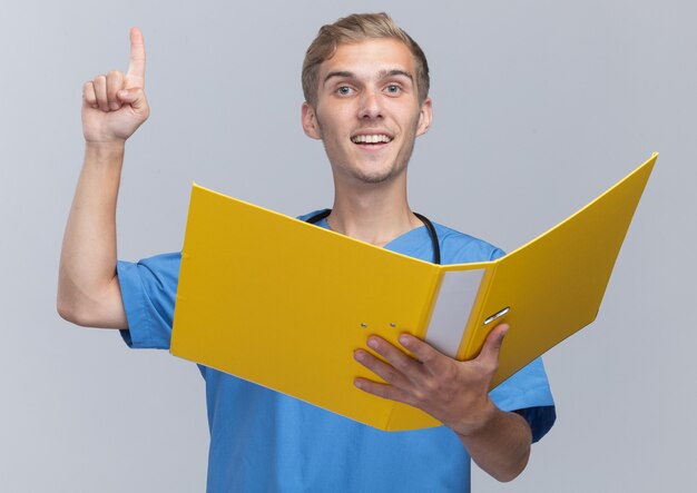 Impressionato giovane medico maschio che indossa l'uniforme del medico con lo stetoscopio che tiene i punti della cartella in alto isolato sul muro bianco