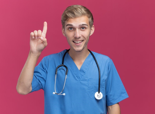 Impressionato giovane medico maschio che indossa l'uniforme del medico con lo stetoscopio punta verso l'alto isolato sulla parete rosa