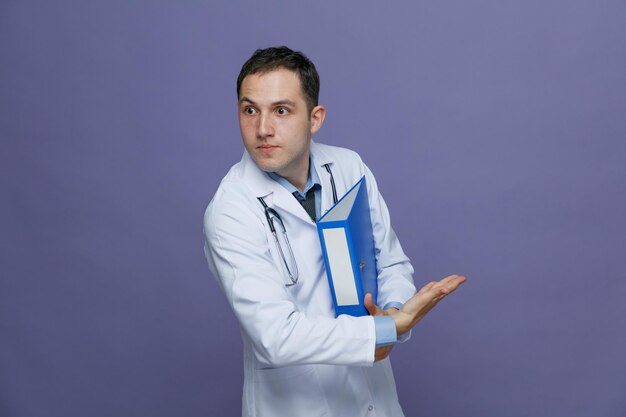 Impressionato giovane medico maschio che indossa un accappatoio medico e uno stetoscopio intorno al collo che tiene la cartella sotto il braccio guardando il lato rivolto a lato con la mano isolata su sfondo viola