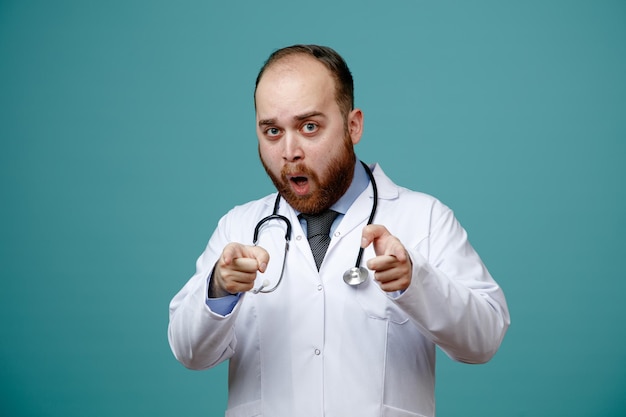 Impressionato giovane medico maschio che indossa un camice medico e uno stetoscopio intorno al collo guardando la fotocamera facendoti un gesto isolato su sfondo blu