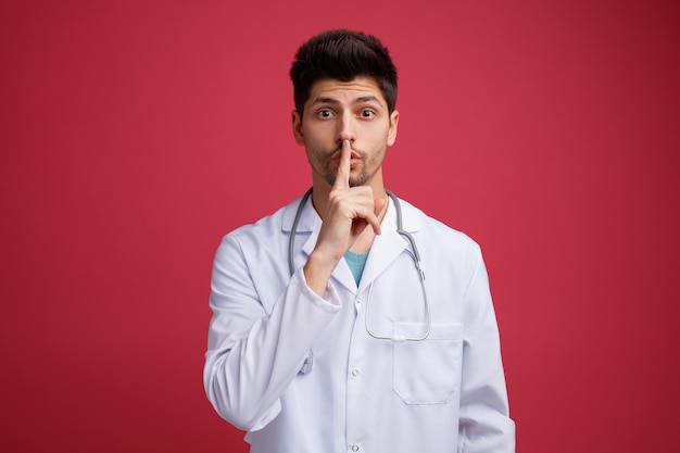 Impressionato giovane medico maschio che indossa uniforme medica e stetoscopio intorno al collo guardando la fotocamera che mostra un gesto di silenzio isolato su sfondo rosso