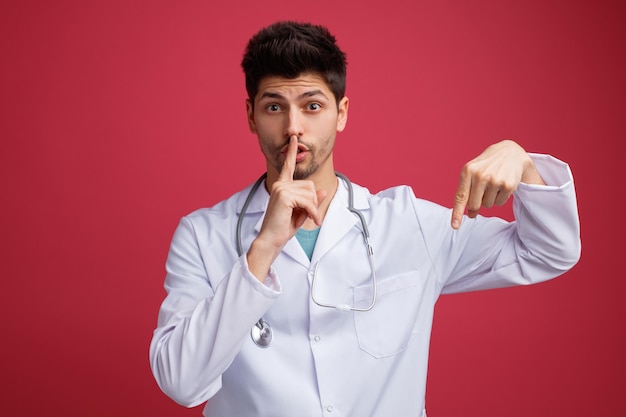 Impressionato giovane medico maschio che indossa uniforme medica e stetoscopio intorno al collo guardando la telecamera rivolta verso il basso che mostra il gesto del silenzio isolato su sfondo rosso