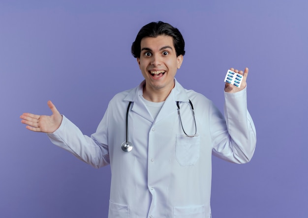 Impressionato giovane medico maschio che indossa veste medica e stetoscopio che mostra il pacchetto di capsule che mostra la mano vuota isolata sulla parete viola