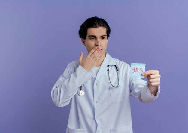 Impressionato giovane medico maschio che indossa veste medica e stetoscopio che mostra sì nota guardandolo tenendo la mano sulla bocca isolata sul muro viola con spazio di copia