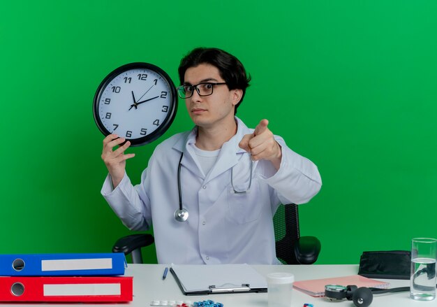 Impressionato giovane medico maschio che indossa veste medica e stetoscopio con gli occhiali seduto alla scrivania con strumenti medici che tengono l'orologio che osserva e che indica isolato sulla parete verde